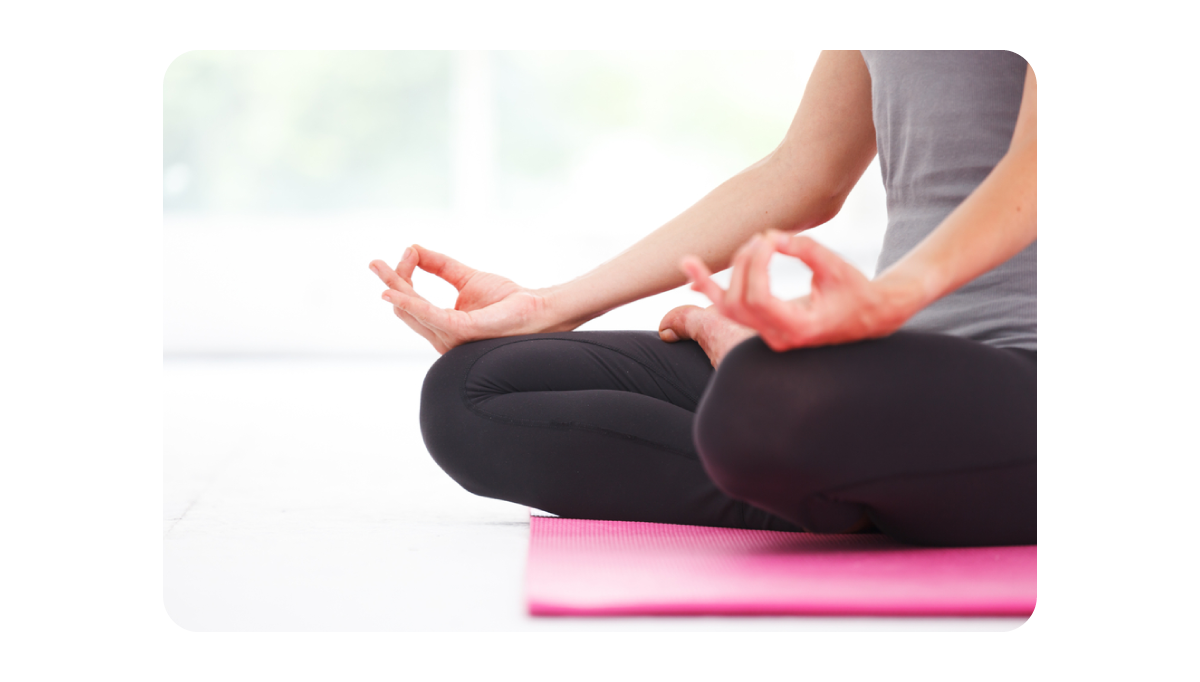 Close-up photo of a person sitting cross-legged on a pink yoga mat, practicing meditation with hands resting on knees in a mudra gesture, wearing black leggings and a grey tank top.png