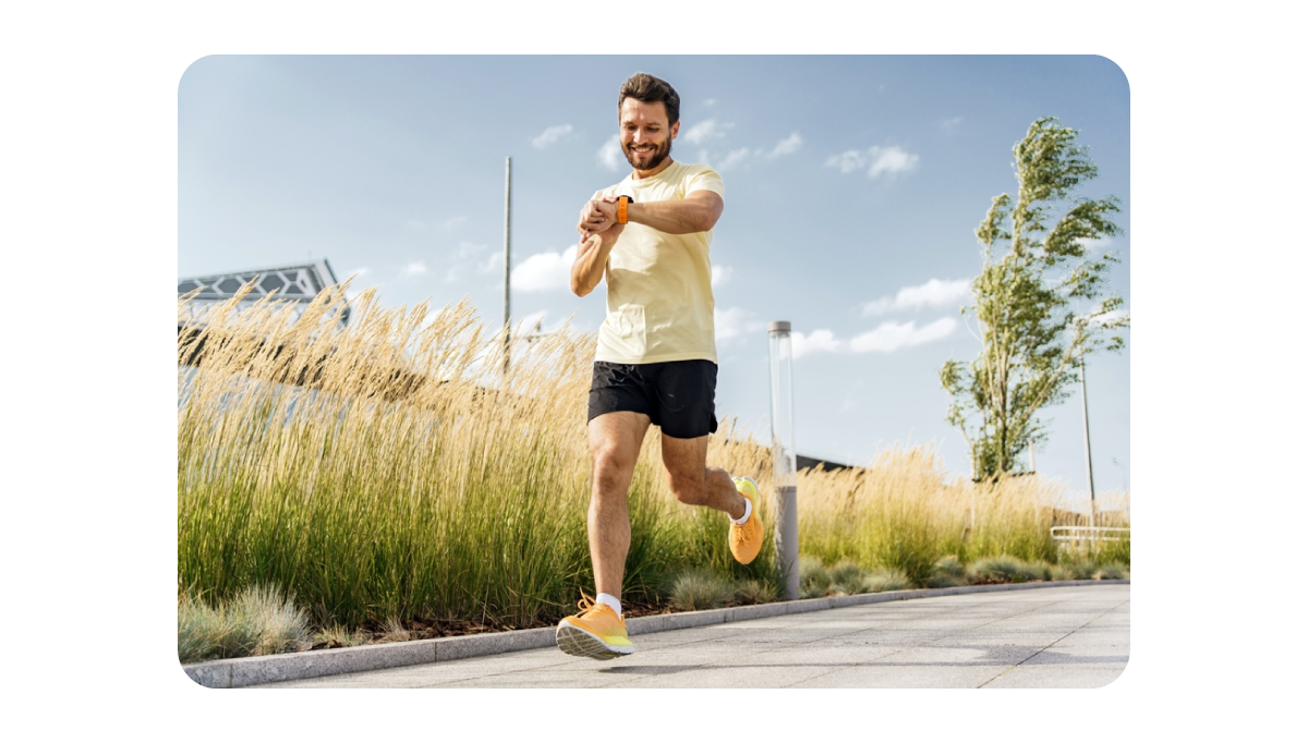 Smiling man jogging outdoors on a paved path, wearing a yellow t-shirt and black shorts, looking at a smartwatch on his wrist, with tall grass and trees in the background on a sunny day.png