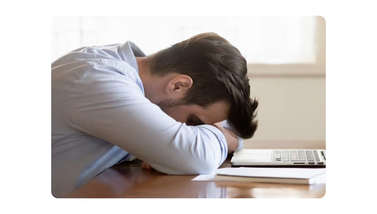 A tired employee napping at his desk with his head resting on folded arms near a laptop.png
